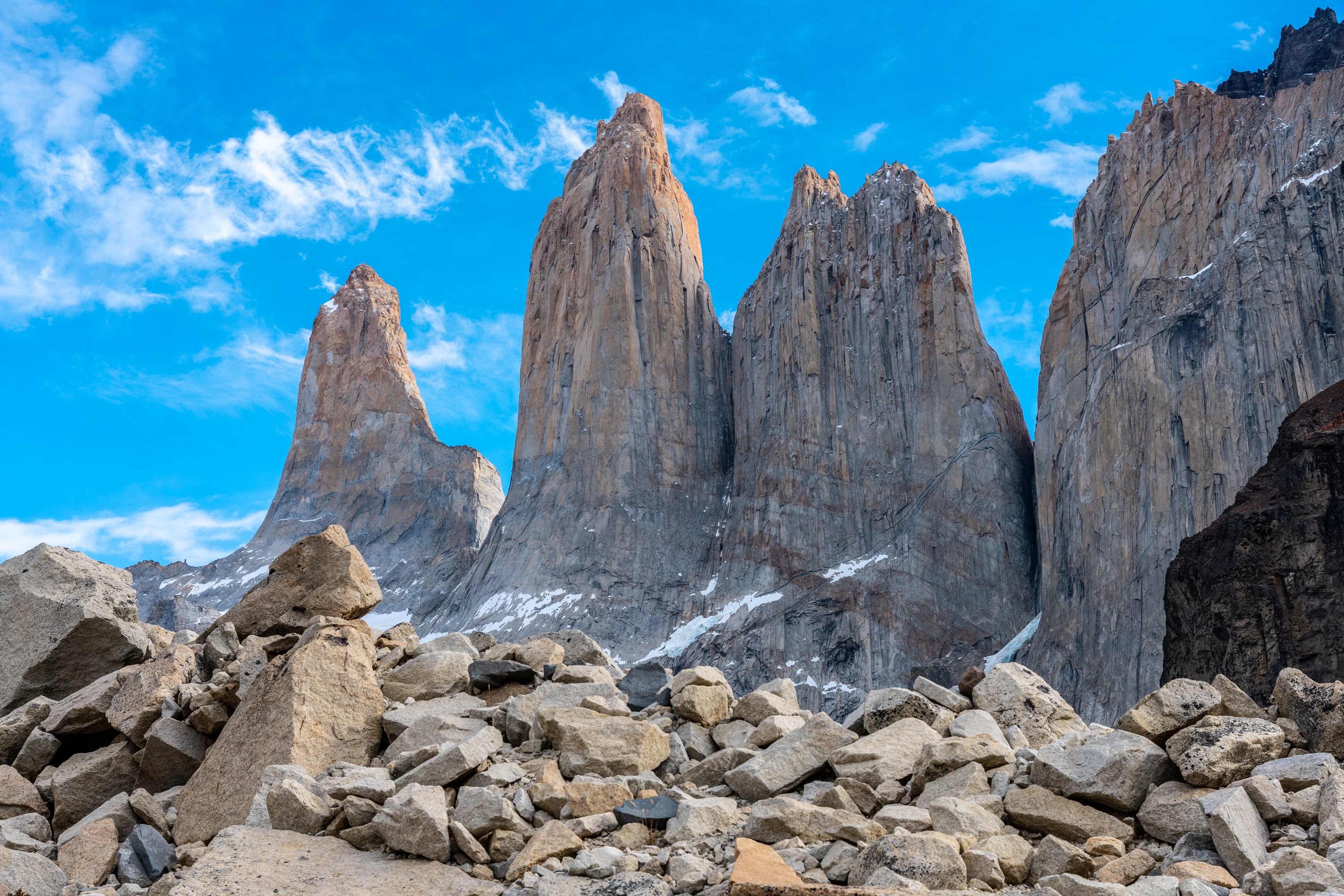 Panoramic view of the Torres del Paine from the Base Torres Viewpoint, featuring three towering granite spires rising above a glacial lake with turquoise waters; surrounded by rugged Patagonian terrain, rocky slopes, and dramatic skies.